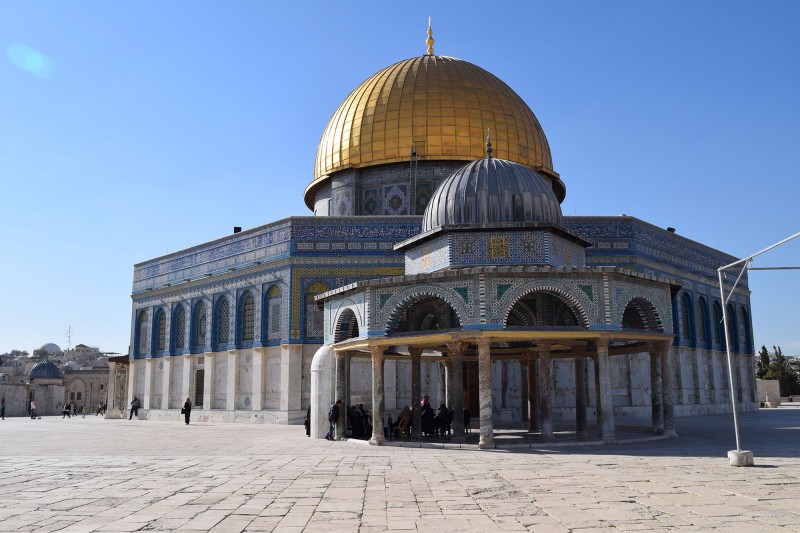 The Dome of the Rock in full view against a clear, blue sky. The richly coloured tiles and golden dome shine brightly in the sun while people gather outside. 